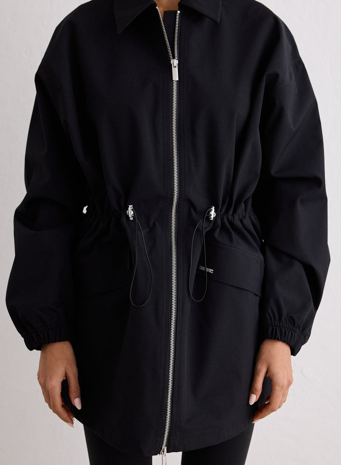 Rajattu sisäkuva mallista, yllään musta Waterproof Zip Jacket. Takissa on hopeinen täyspitkä vetoketju, joka ulottuu takin helmasta kaulukseen asti, ja hopeinen vetoketjun vedin on selkeästi näkyvissä. Hopeisilla metallipäillä varustettu kiristysnyörillinen vyötärö korostaa takkia, ja mallin käsi vaaleanpunaisilla manikyyrikynsillä lepää kuvan oikeassa alakulmassa. [S-170cm]