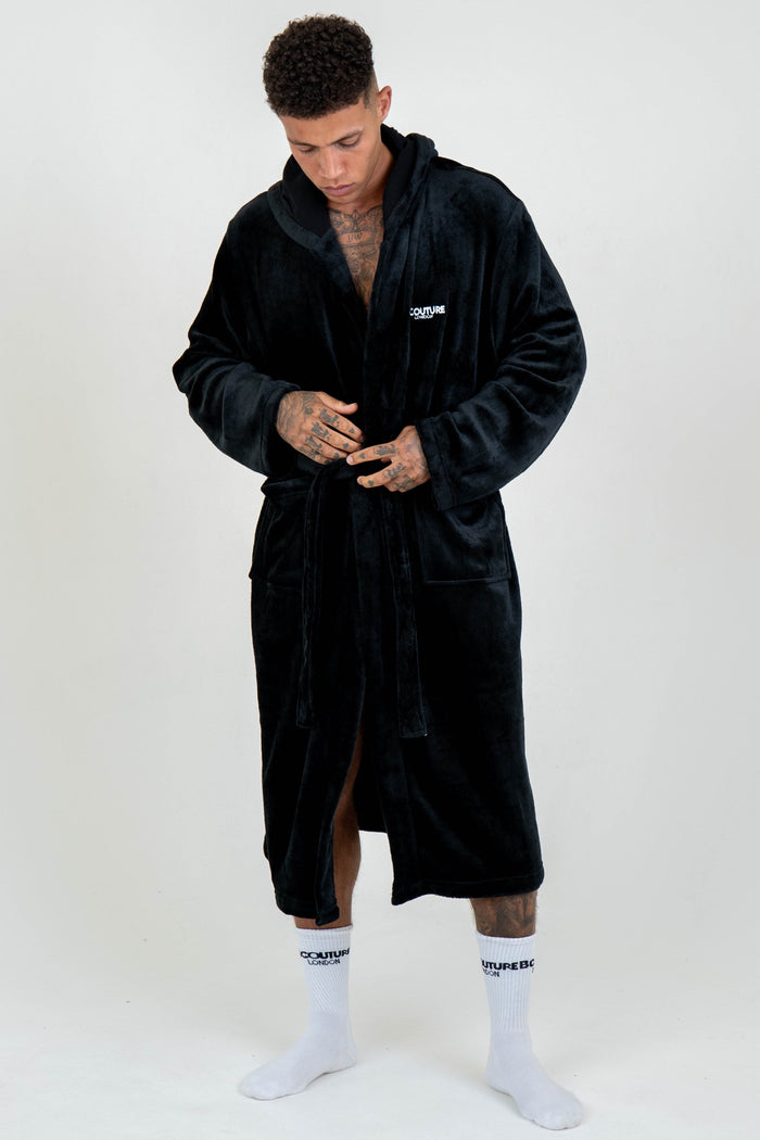 A person stands on a white background wearing the Earnshaw Dressing Gown - Black, a hooded men's luxury robe with a belt and small left chest logo, revealing tattoos on their chest, neck, and hands, paired with white socks.
