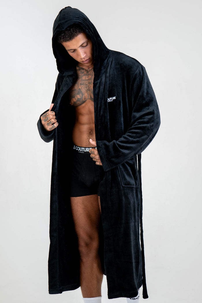 A man wearing the Earnshaw Dressing Gown - Black with the front open, showing his tattooed chest and black shorts, stands against a plain white background and looks down.