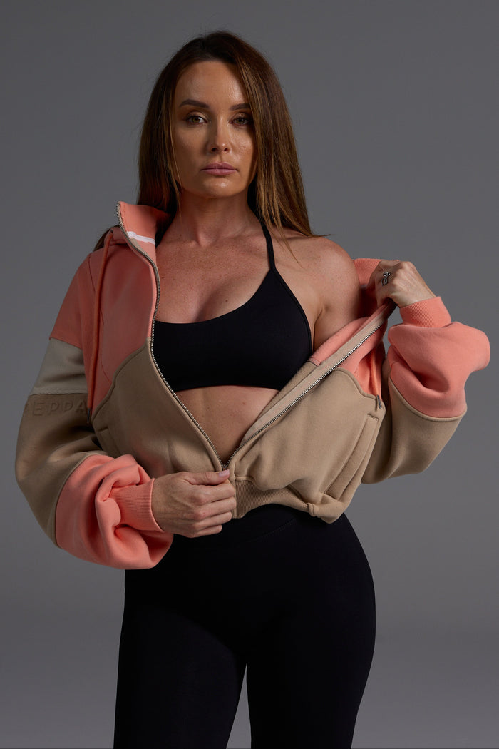 Coral and desert fleece hoodie worn unzipped over black sports bra, demonstrating versatile layering and gym-to-street styling