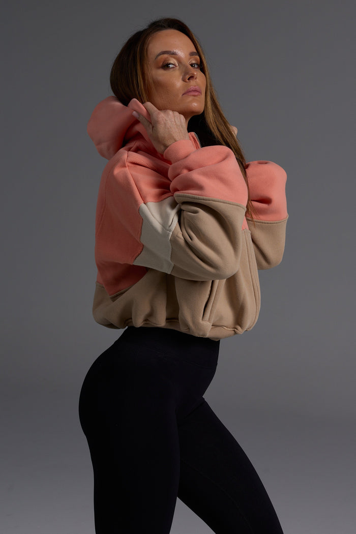 Woman wearing oversized coral and desert colorblock fleece hoodie with black leggings, showing the structured fit and premium heavyweight fabric