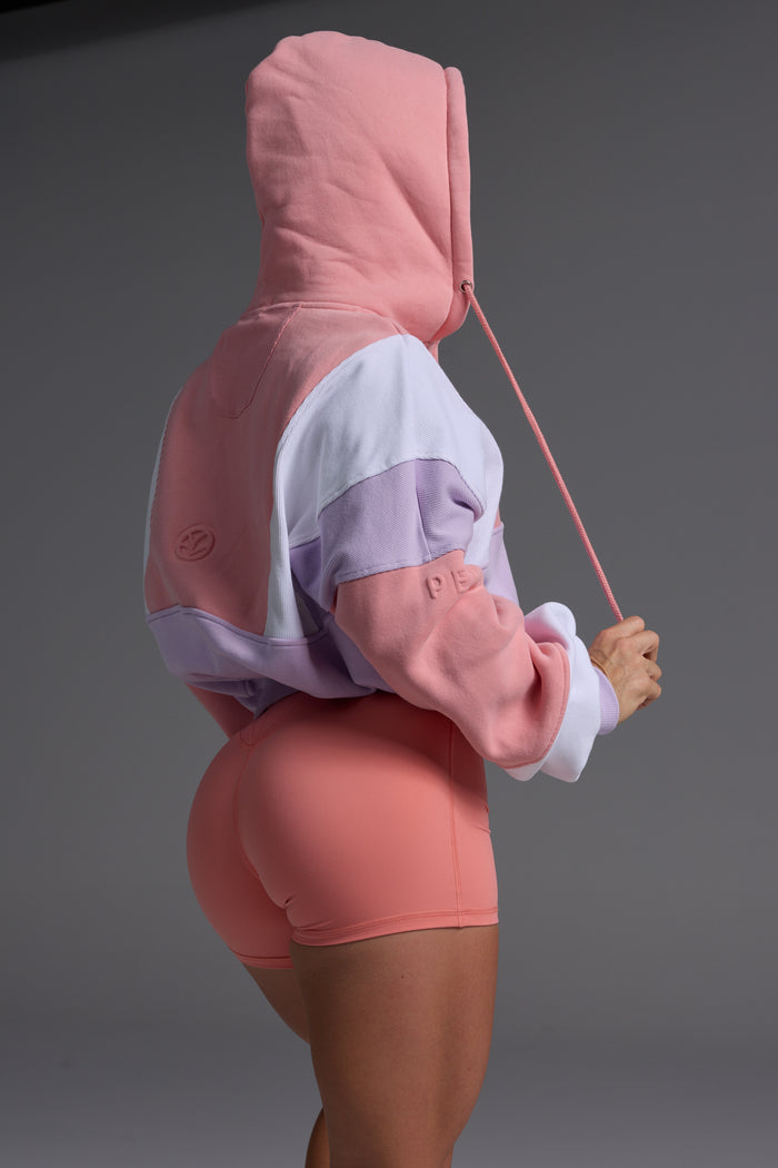 Side profile of woman in peach, lilac and white oversized fleece hoodie with coral bike shorts against grey studio background