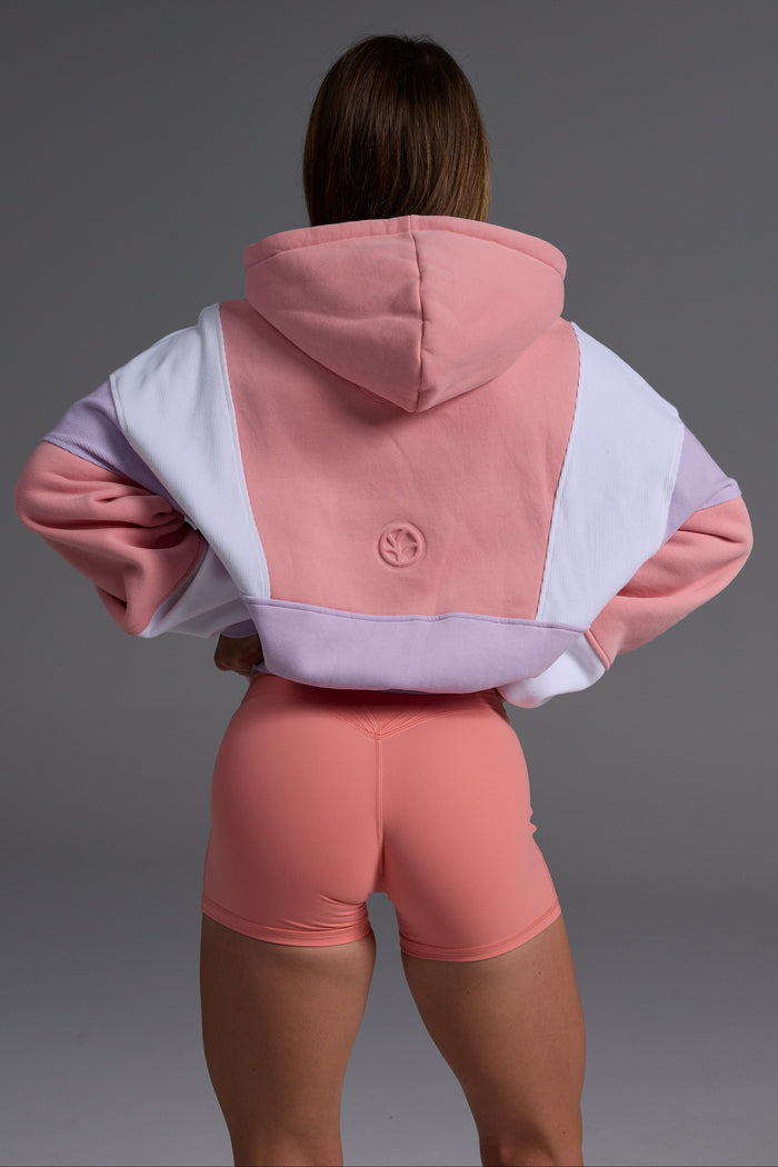 Woman in peach, lilac and white oversized fleece hoodie with coral bike shorts in confident pose against grey background