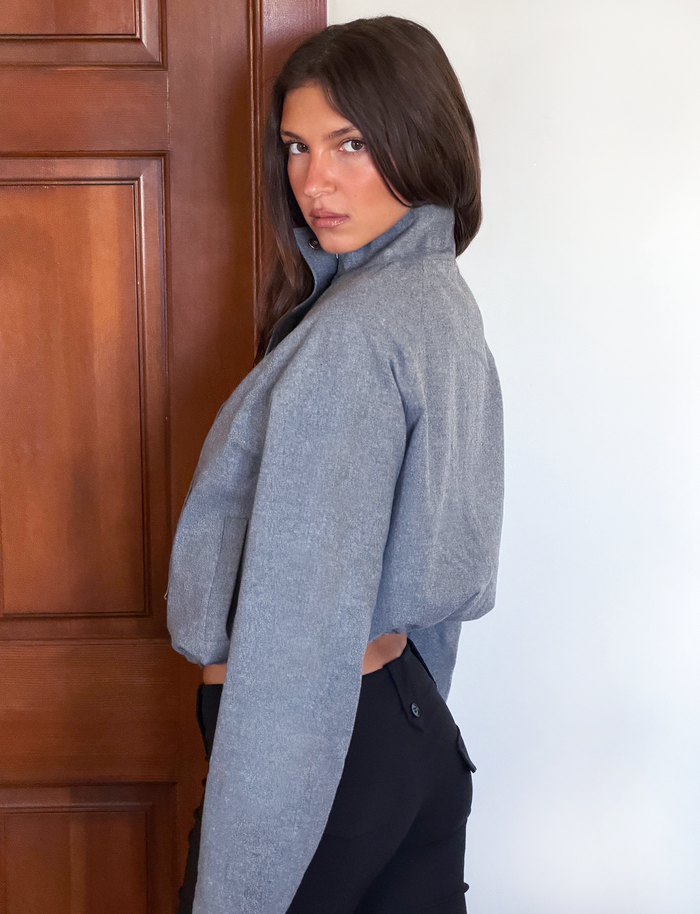 A woman with long brown hair stands sideways, looking over her shoulder at the camera. She wears the TIGERMIST LENNEX JACKET in Heather Charcoal and black pants, with a wooden door and white wall behind her.