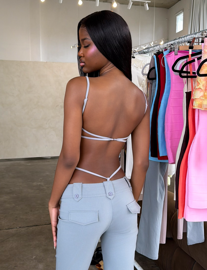 A woman with long, straight hair models the TIGERMIST MINERVA BODYSUIT in frost grey and light blue pants, standing by a clothing rack with colorful pieces in a modern minimalist room.