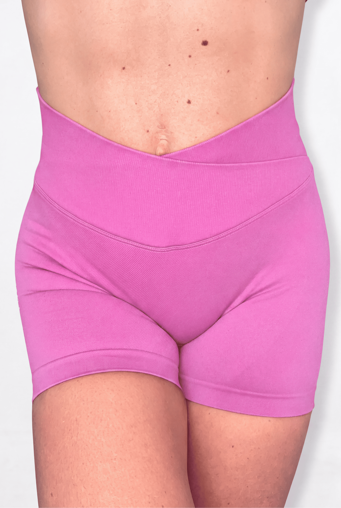 A close-up of a model wearing high-waisted, crossover-front athletic shorts in a solid light pink colour, shown from the waist to the upper thighs against a plain white background.