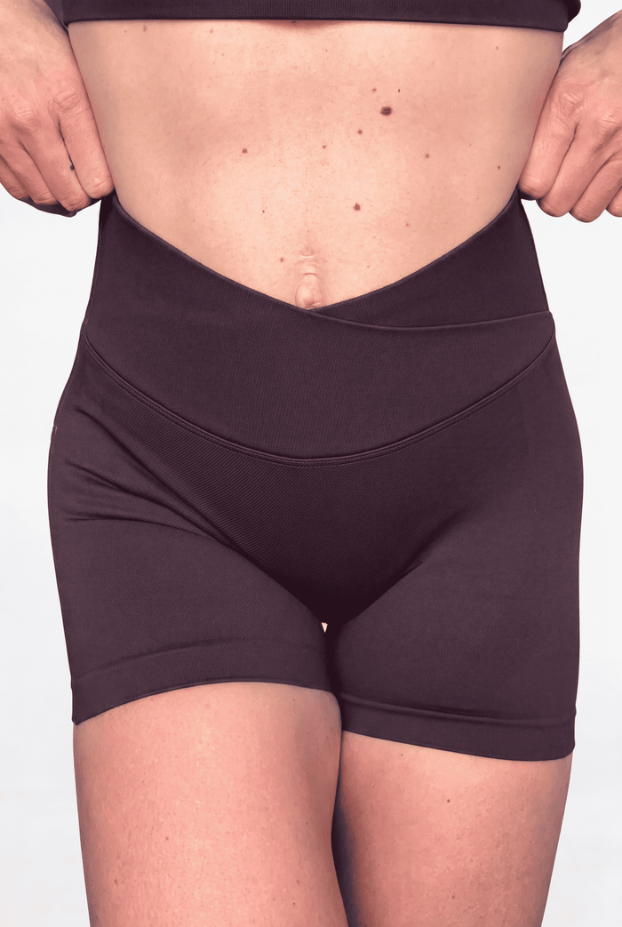 A close-up of a model wearing high-waisted, crossover-front athletic shorts in a chocolate brown colour, shown from the waist to the upper thighs against a plain white background.