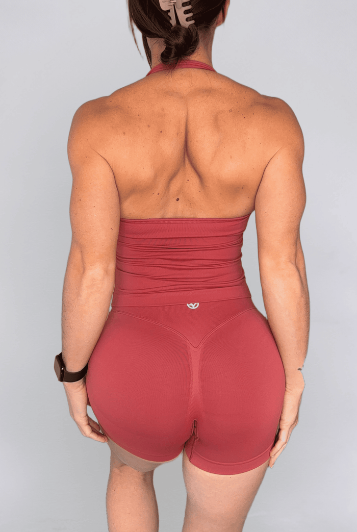 Rear view of a woman wearing a terracotta high-waisted athletic set consisting of a strapless tank top and matching high-waisted shorts, on a light gray background.