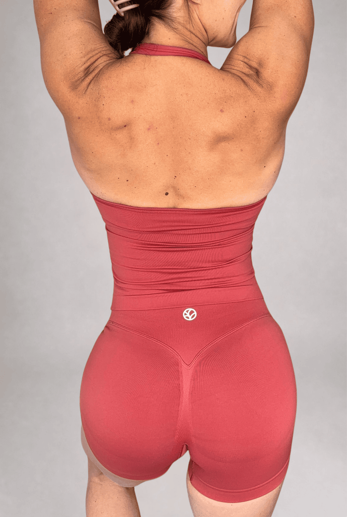 Rear view of a woman wearing a terracotta athletic halter tank top with matching shorts, featuring a small white circular logo on the lower back, set against a plain gray studio background.